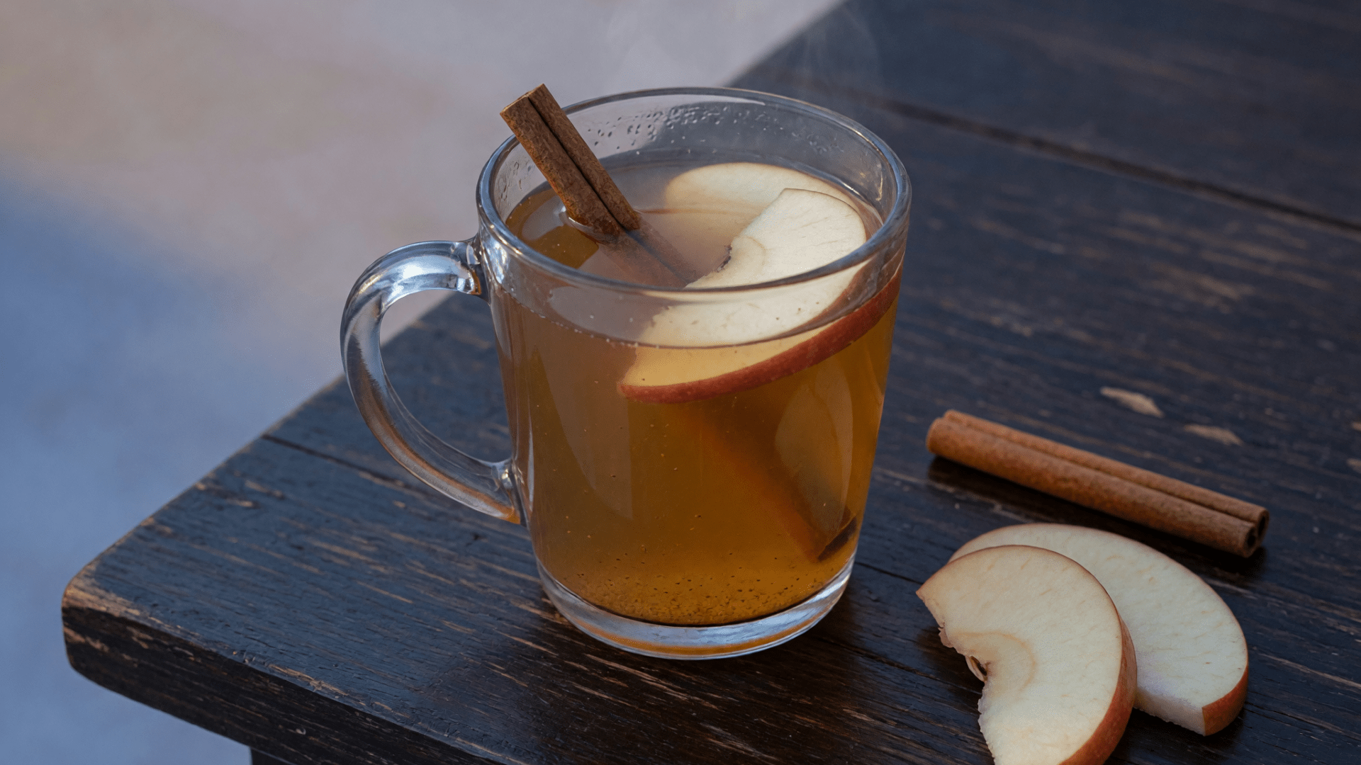 Tasse en verre de boisson ambrée aux épices, cannelle et tranches de pomme sur table sombre.
