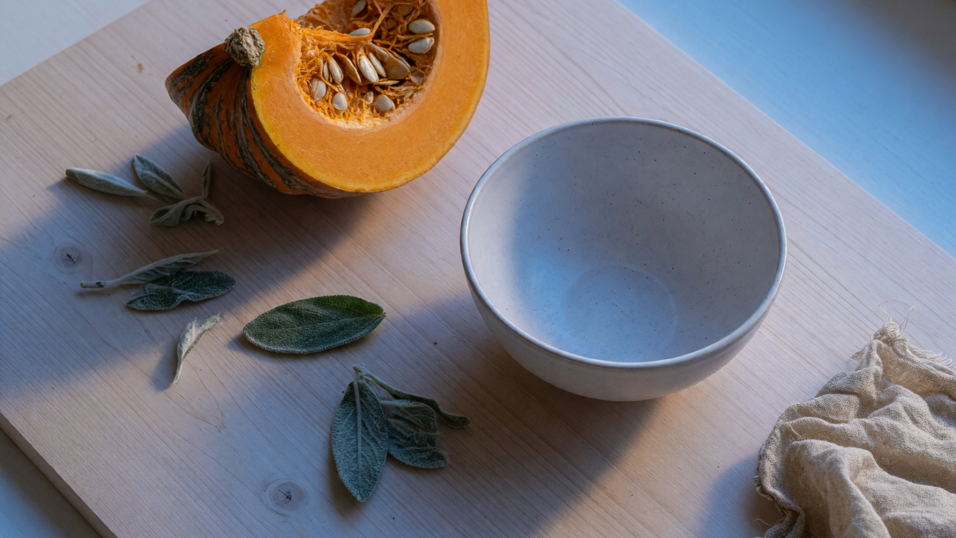 Une citrouille coupée, des feuilles de sauge et un bol en céramique reposent sur une planche claire.