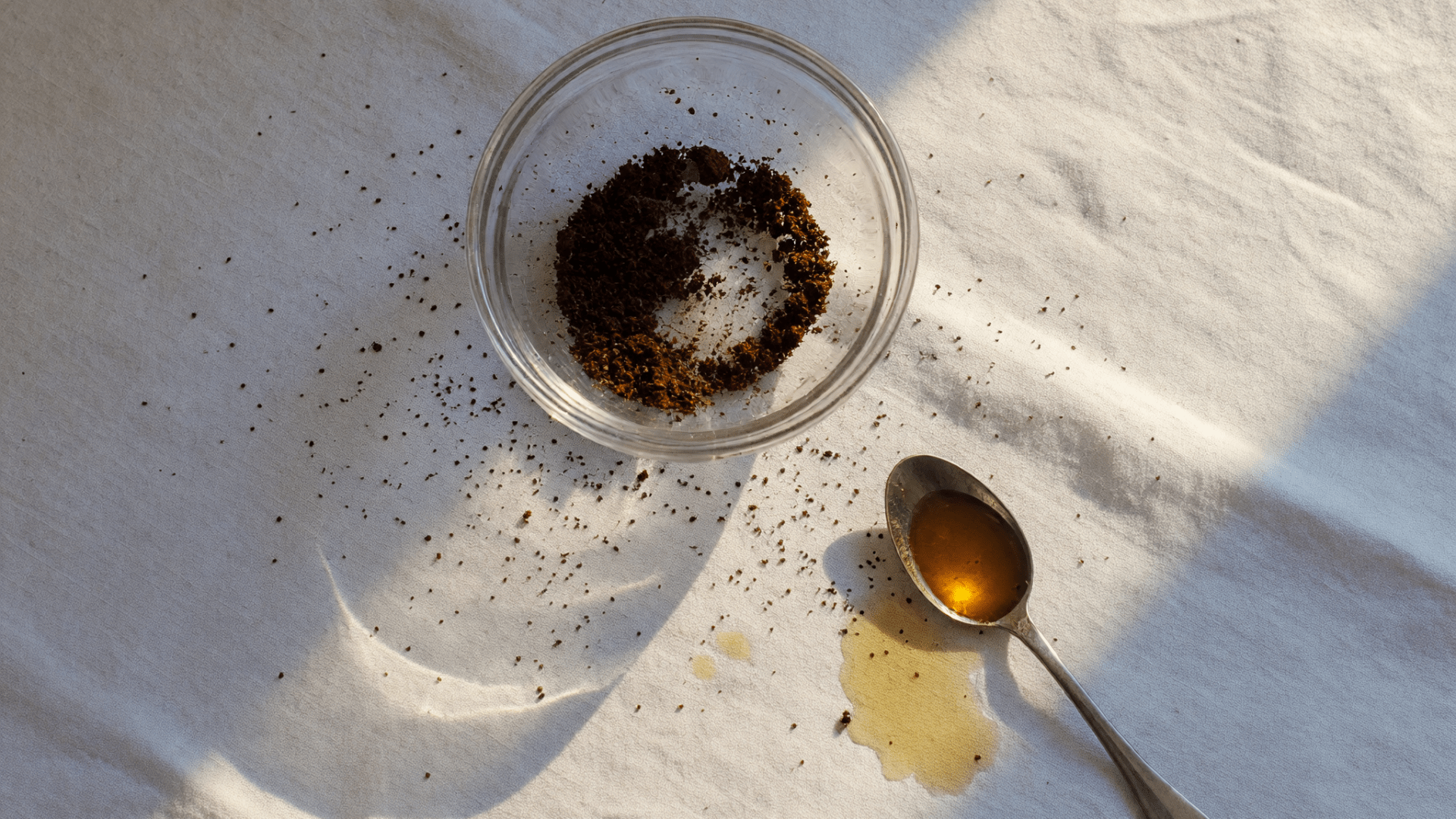 Marc de café dans un bol en verre, cuillère d’huile et lumière matinale sur linge sable.
