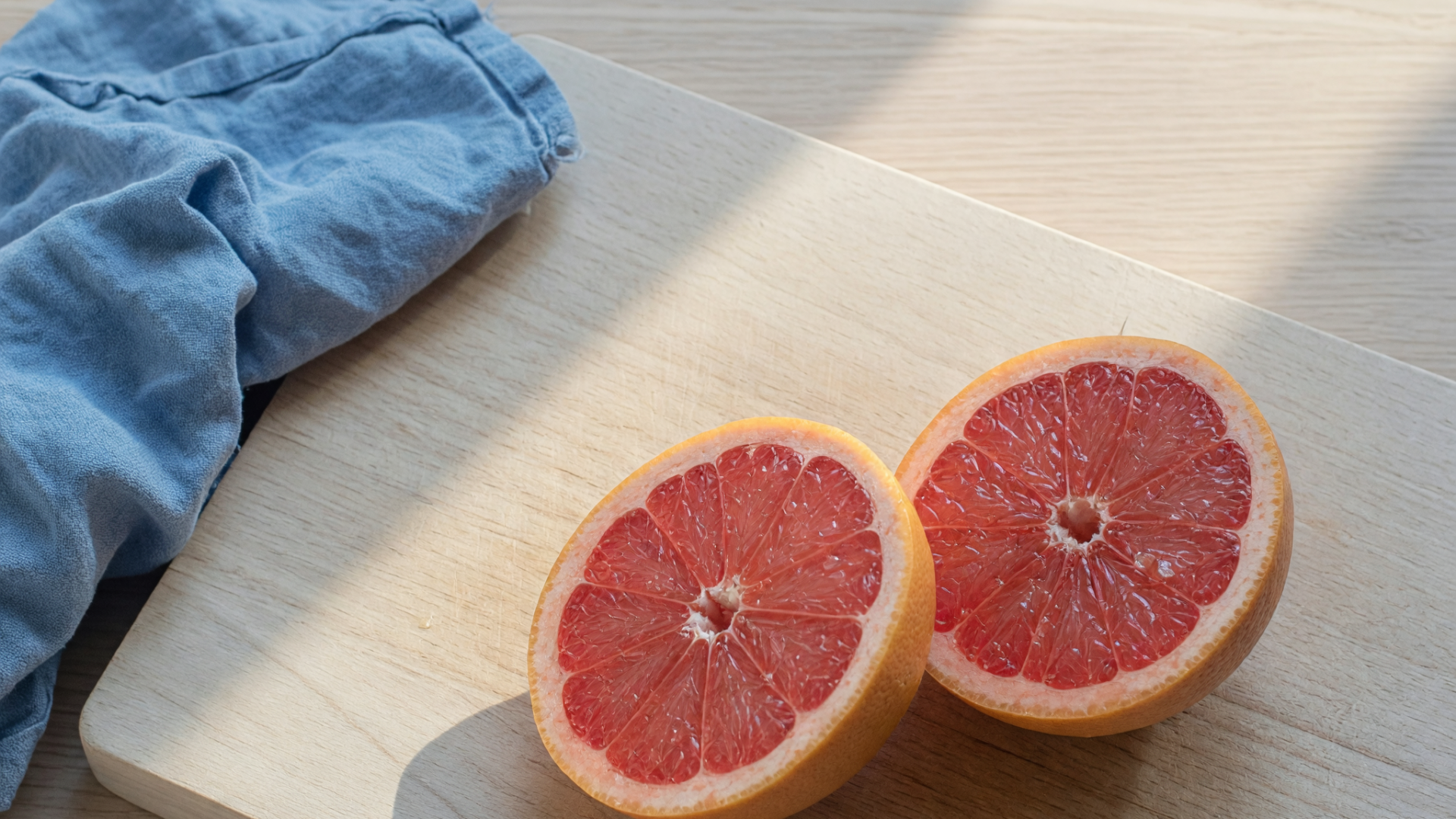 Deux moitiés de pamplemousse reposent sur une planche en bois clair, avec un tissu bleu dans un coin.