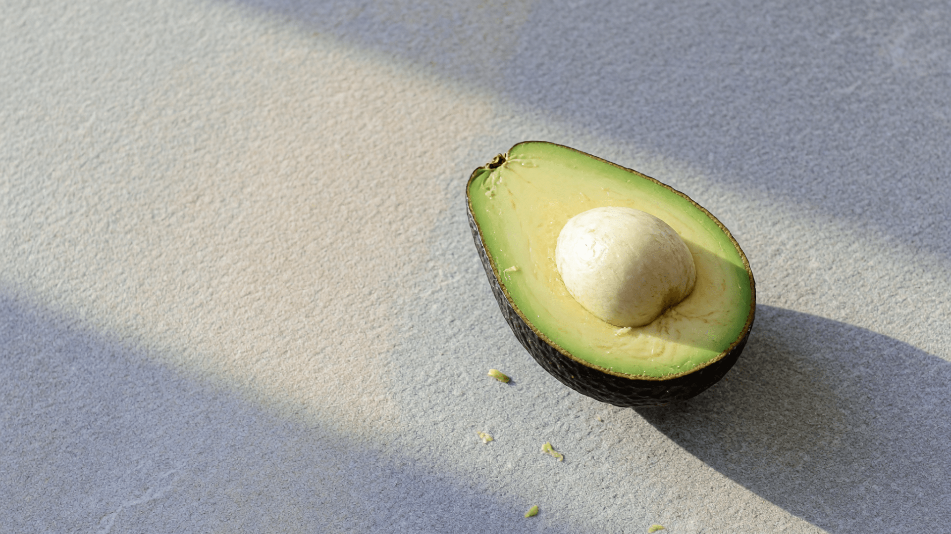 Une moitié d’avocat avec son noyau repose sur une surface grise, éclairée latéralement par le soleil.