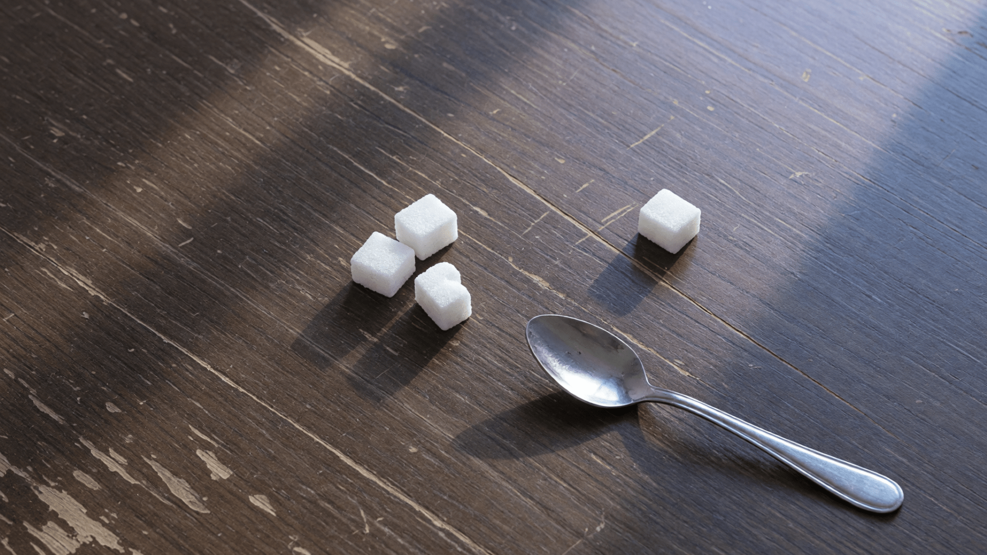 Quatre morceaux de sucre et une petite cuillère en métal sont posés sur une table en bois sombre.