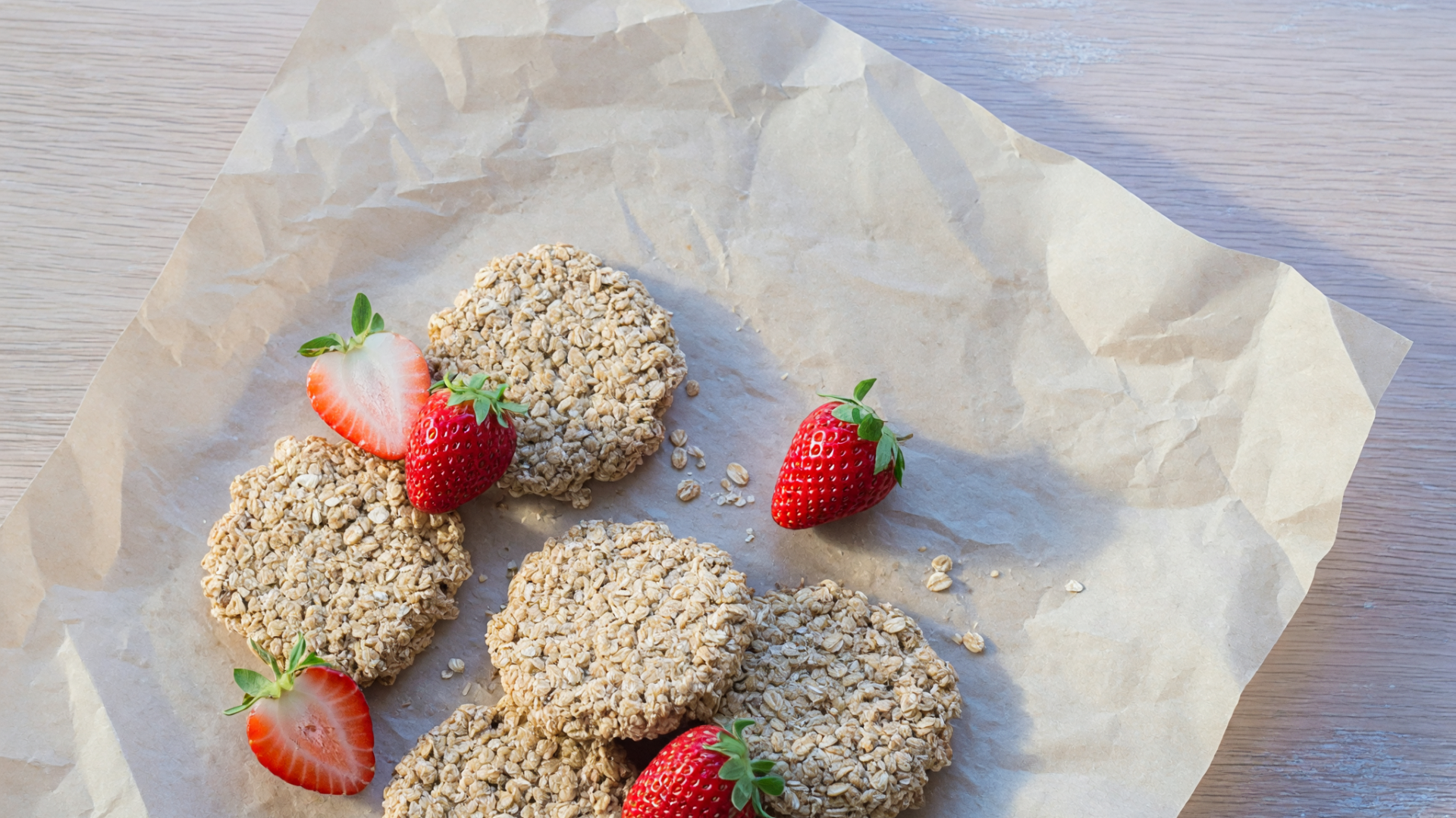 Des biscuits à l'avoine et quelques fraises sont posés sur du papier cuisson, sur une table claire.