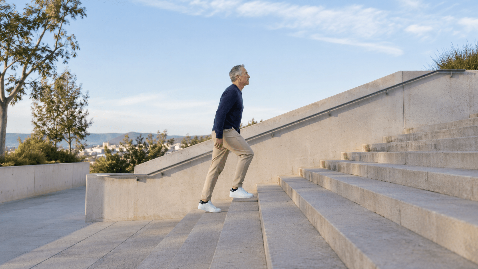 Un homme aux cheveux gris, en pull marine, monte un escalier de béton à l'extérieur.