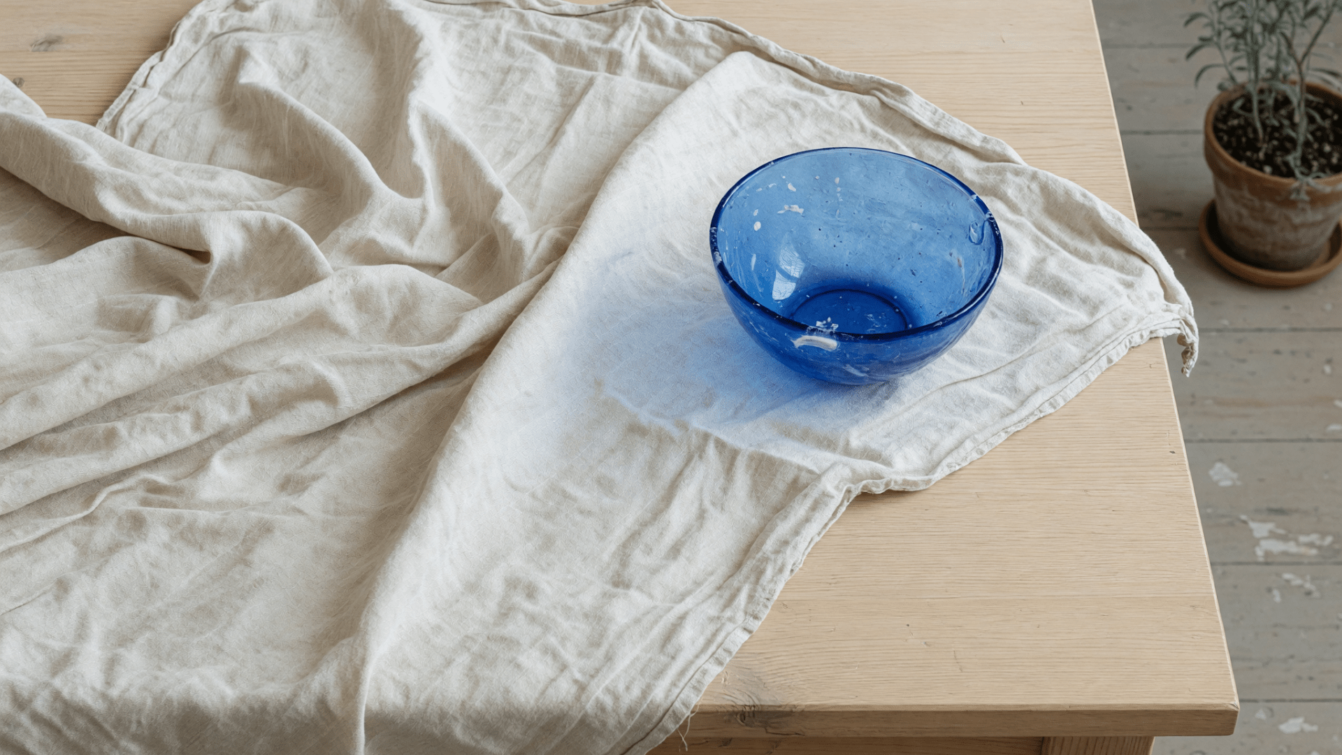 Un linge beige froissé et un bol en verre bleu sont posés sur une table en bois, près d'une plante.