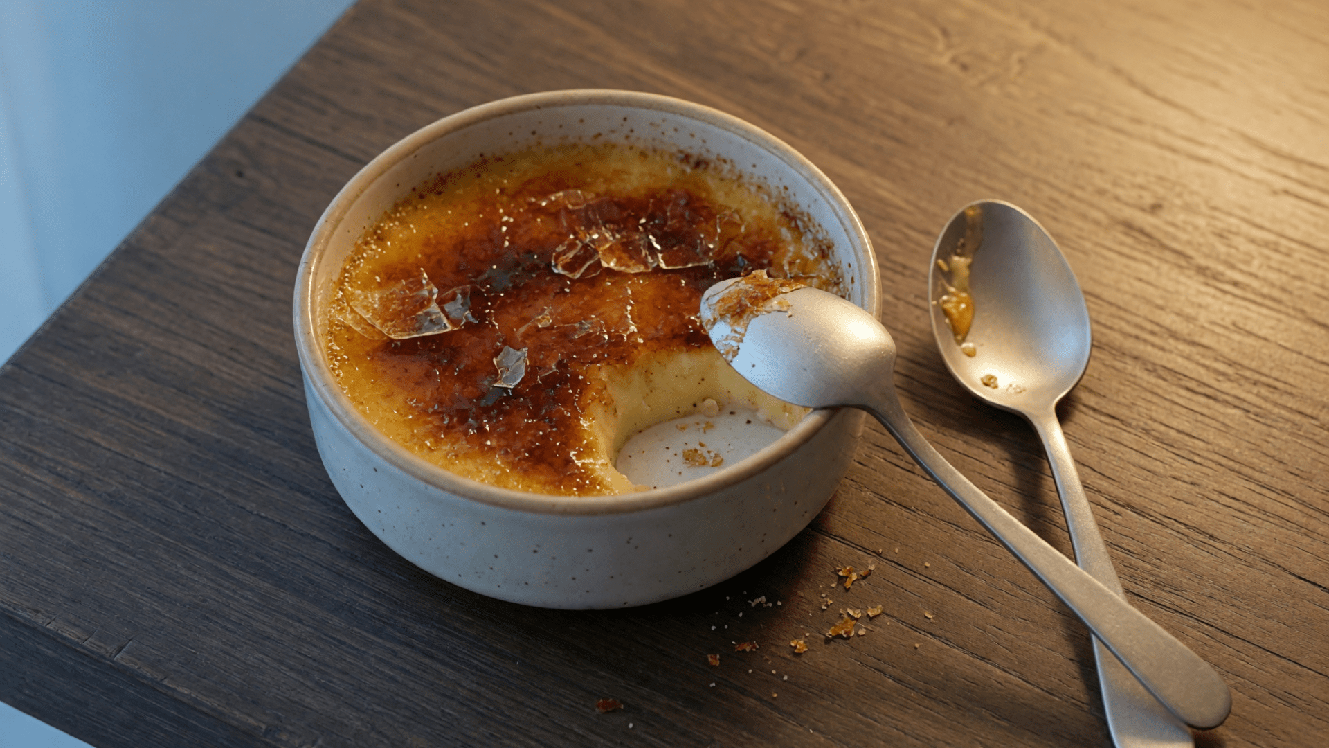 Un ramequin de crème brûlée entamée et deux cuillères sont posés sur une table en bois foncé.
