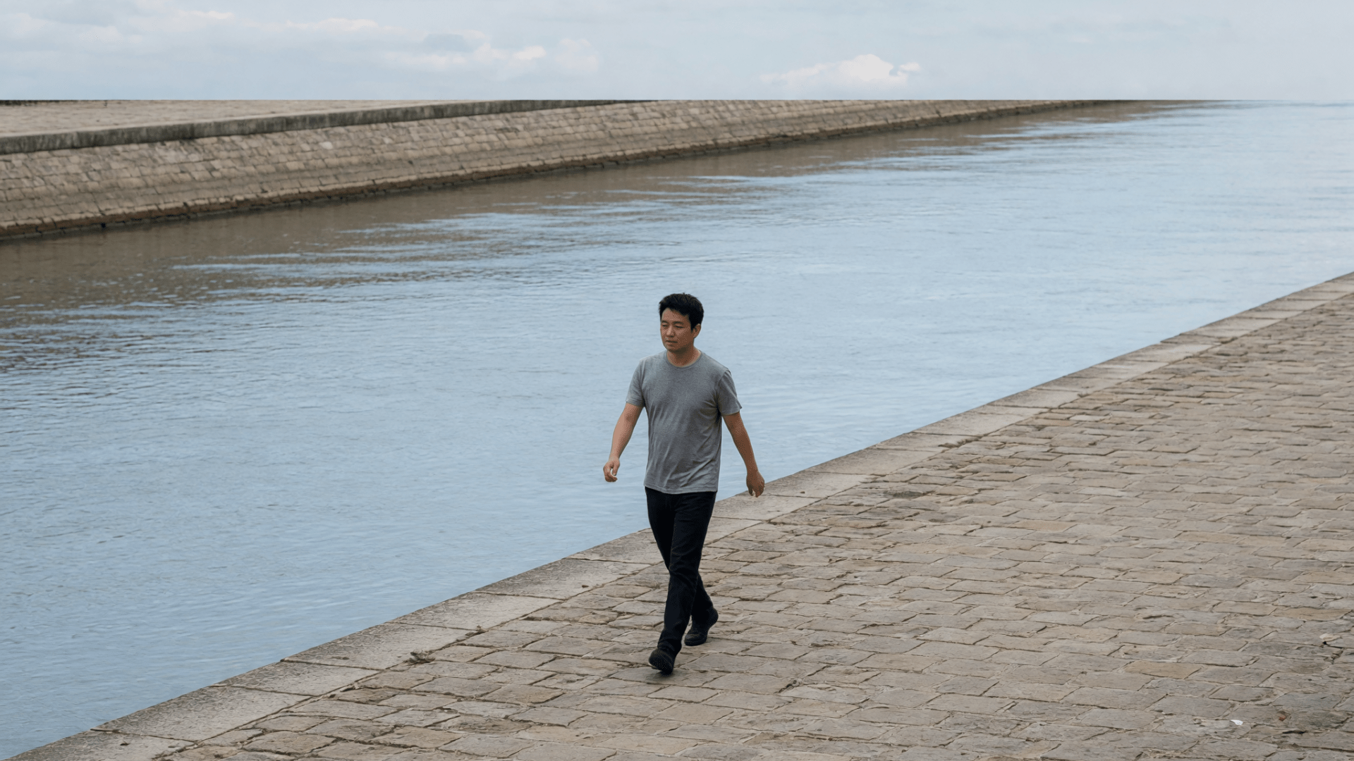 Un homme en t-shirt gris marche sur un quai de pierre, le fleuve derrière lui.