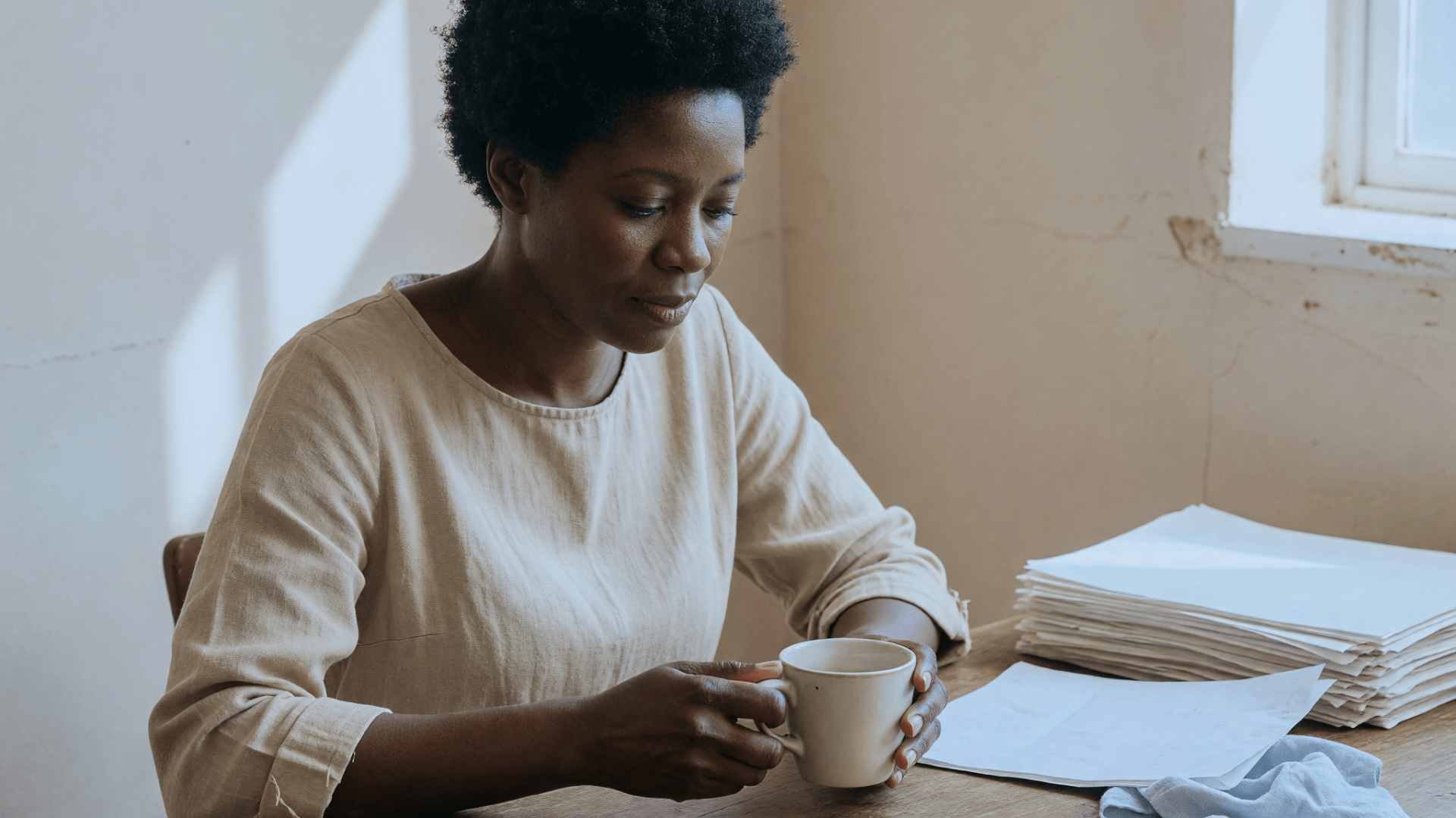 Une femme aux cheveux courts, en pull clair, tient une tasse à deux mains, à un bureau en bois.