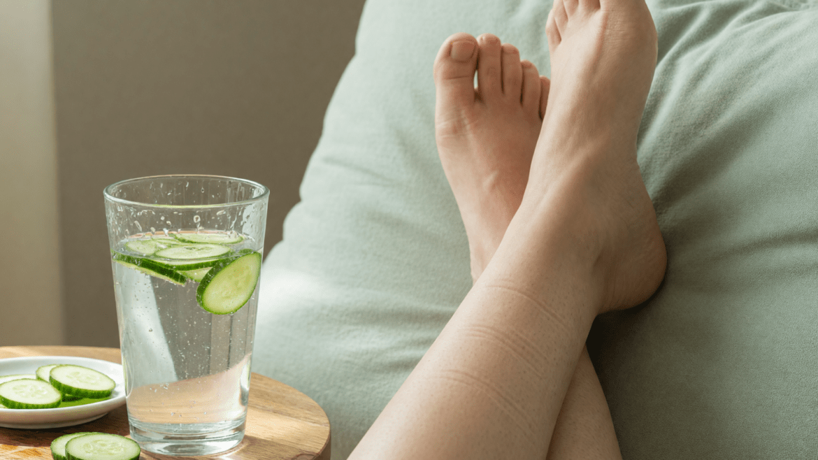 Las piernas están en alto, al lado de un vaso de agua con pepino.