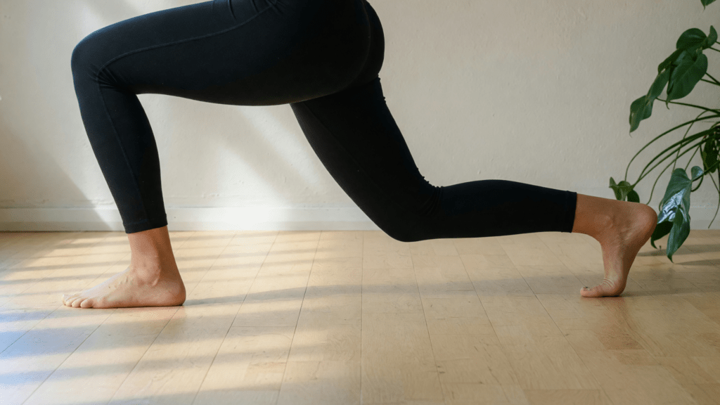 Uma pessoa faz um afundo no chão de madeira, de legging preta.