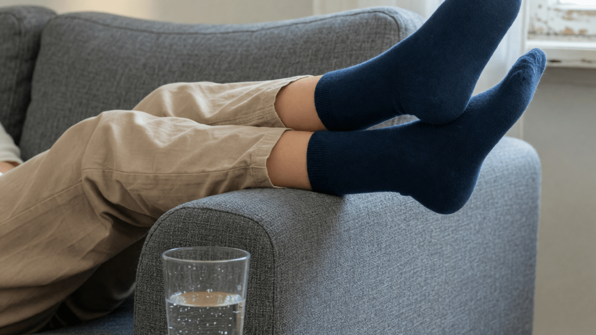Beine in dunkelblauen Socken liegen hoch auf der Armlehne eines Sofas, neben einem Glas Wasser.