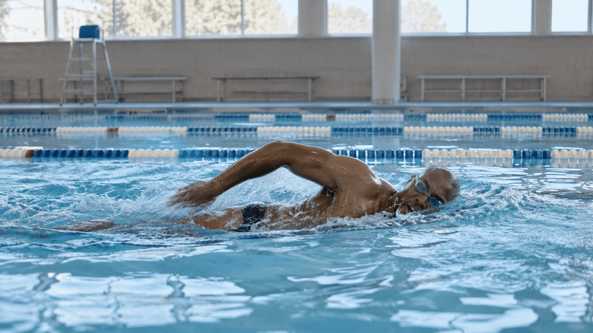 Un hombre nada a crol en una piscina cubierta, con gafas y bañador oscuro.
