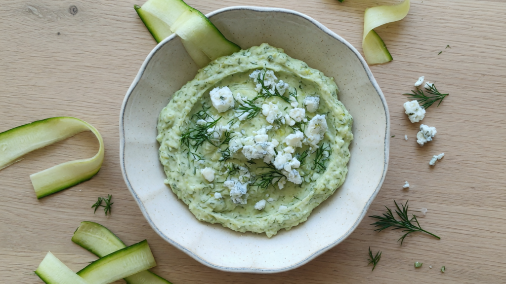 Un bol de dip vert pâle, garni de fromage blanc et d'aneth, entouré de rubans de courgette.