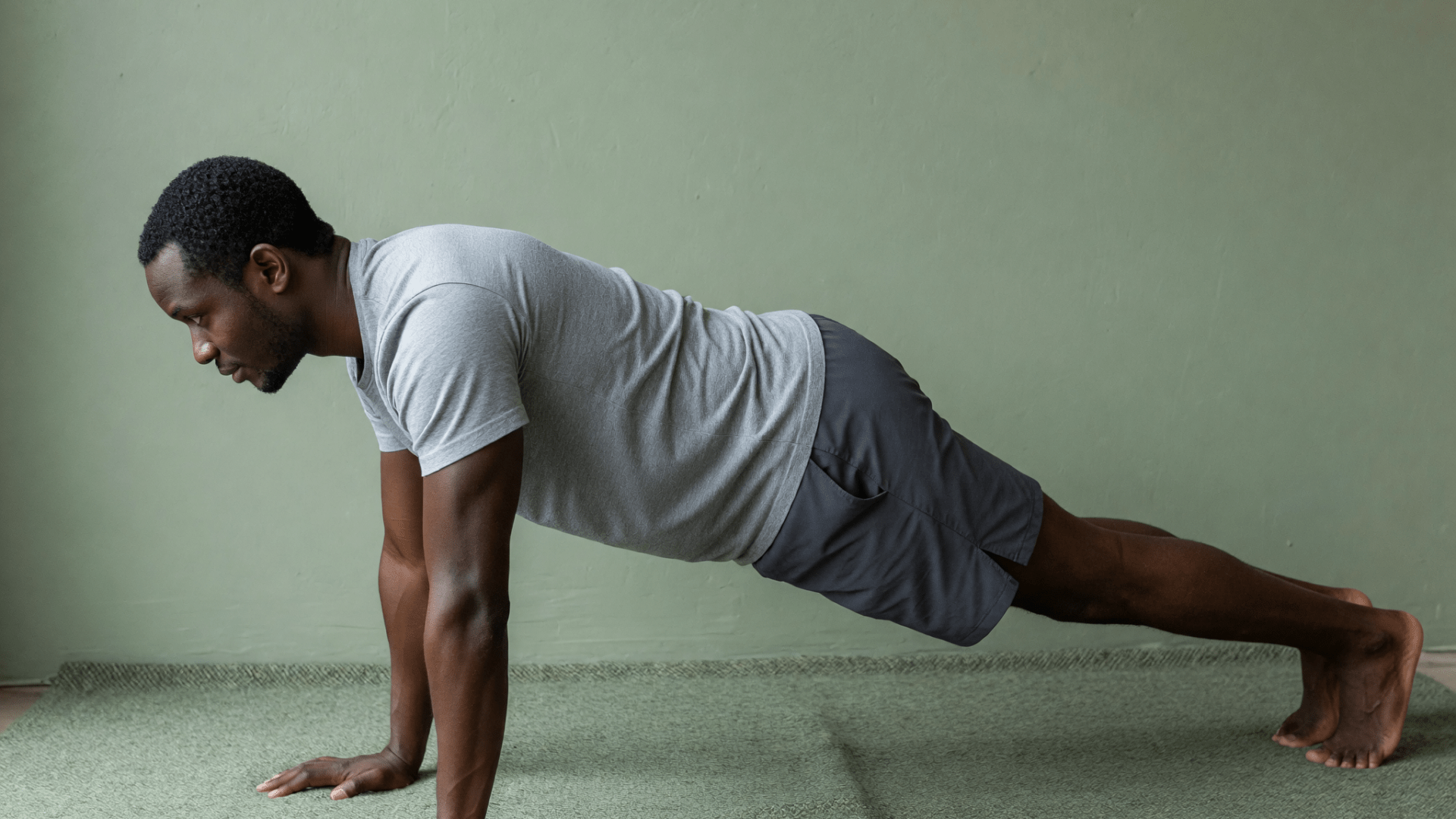 Un homme en t-shirt gris tient une planche sur les mains, devant un mur vert.