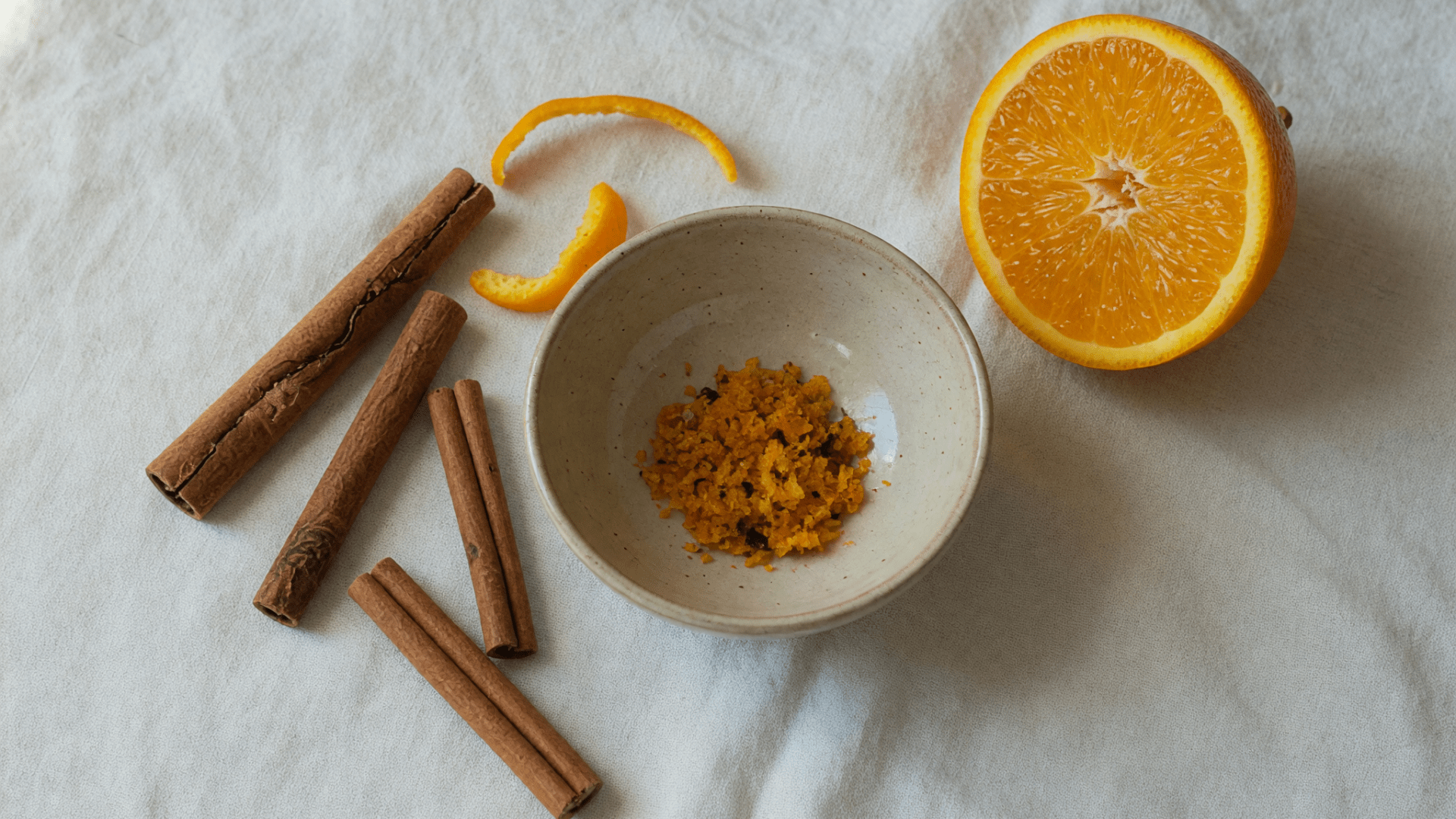 Des bâtons de cannelle, une demi-orange et un bol de zeste râpé sur un tissu clair.