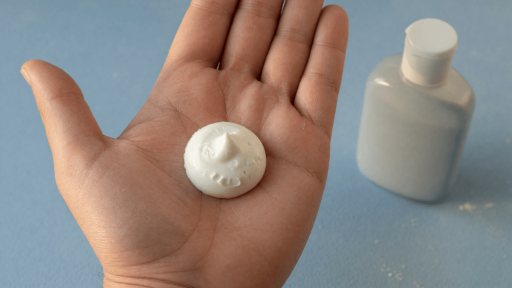 Gros plan d’une paume ouverte contenant une noisette de crème blanche, avec un flacon sans étiquette en arrière-plan sur une surface bleue.