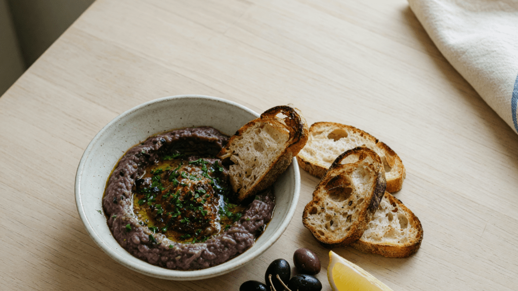 Un bol de caviar d'aubergines, du pain grillé, des olives et un citron, sur une table en bois.