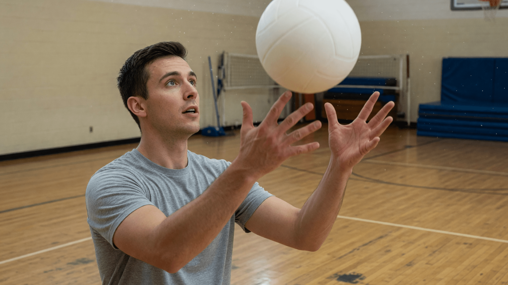 Un homme en t-shirt gris joue au volley dans un gymnase, avec un ballon blanc.